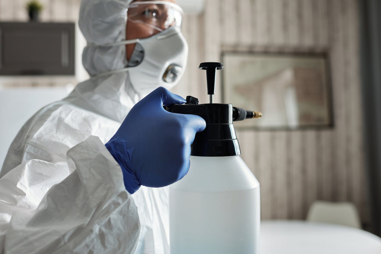A person in protective gear with gloves and face mask using a spray bottle for disinfection indoors.